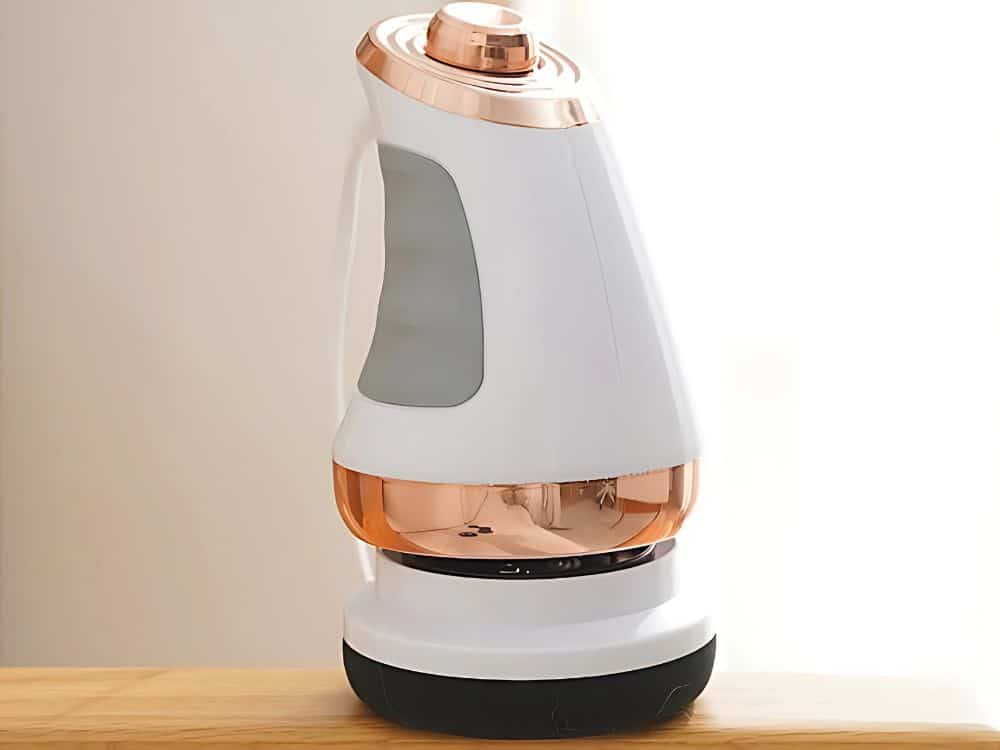 Bílá designová elektrická konvice s měděnými detaily a průhlednou spodní částí, umístěná na dřevěném povrchu s neutrálním pozadím.