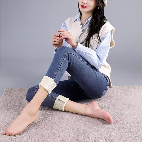 Žena sedící na koberci v bílé vestě a modré košili, oblečená v modrých džínách, bosá a s rukama sepjatýma před sebou.