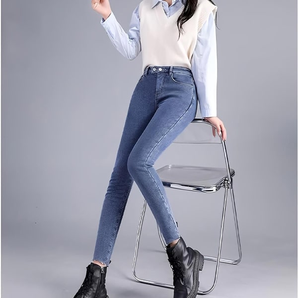 Žena v bílé vestě a světle modrých džínách stojící u skládací kovové židle na šedém pozadí, oblečená v casualním stylu s černými kotníkovými botami.