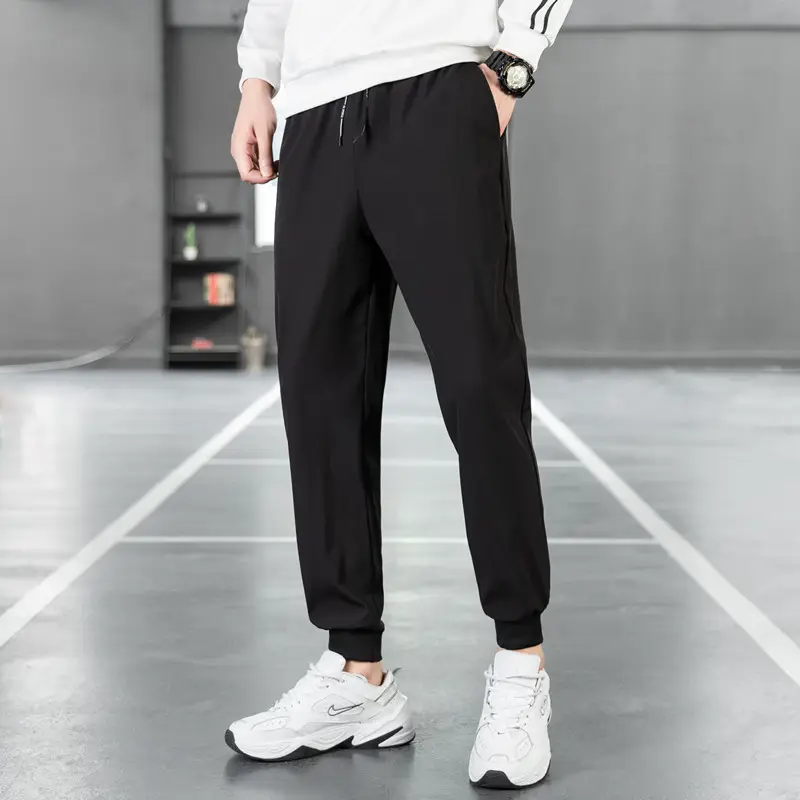 Černé sportovní kalhoty s úzkým střihem, stahovací na bocích, kombinované s bílými nadměrnými tenisky Nike na šedém pozadí.