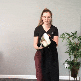 Žena v černých šatech stojí u zdi a drží malou bílou skulpturu, vedle ní je zelená pokojová rostlina v květináči.