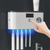 Elektrický držák na zubní kartáčky s UV sterilizací, na kterém je právě zapínán pomocí prstu a který je umístěn v koupelně vedle umyvadla.