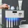 Elektrický držák na zubní kartáčky s UV sterilizací a dávkovačem zubní pasty, umístěný na šedé koupelnové stěně s kosmetickými přípravky a elegantním designem.