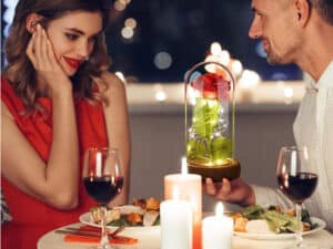 Romantická večera pri sviečkach s červeným vínom, muž drží sklenený kryt s ružou inšpirovanou rozprávkou Kráska a zviera, žena v červených šatách sa usmieva.