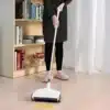 I see a floor sweeper/cleaning device being used by someone wearing black clothing and checkered slippers. The device appears to be white with wheels, and there's a light patch on the floor which suggests it's picking up something while being pushed.