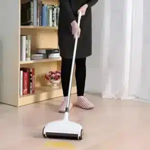 I see a floor sweeper/cleaning device being used by someone wearing black clothing and checkered slippers. The device appears to be white with wheels, and there's a light patch on the floor which suggests it's picking up something while being pushed.