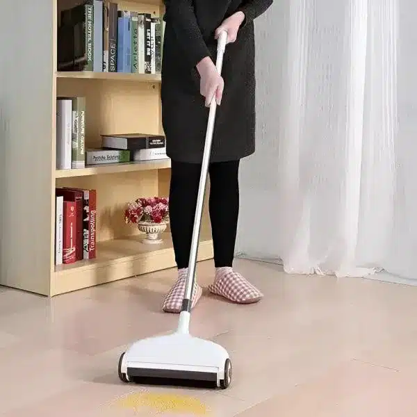 I see a floor sweeper/cleaning device being used by someone wearing black clothing and checkered slippers. The device appears to be white with wheels, and there's a light patch on the floor which suggests it's picking up something while being pushed.
