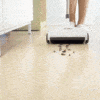 A white cordless floor sweeper cleaning up scattered coffee beans on a light wooden floor, with part of a person's legs visible in the background.