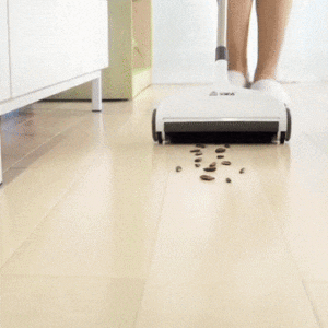 A white cordless floor sweeper cleaning up scattered coffee beans on a light wooden floor, with part of a person's legs visible in the background.