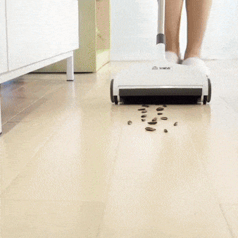 A white cordless floor sweeper cleaning up scattered coffee beans on a light wooden floor, with part of a person's legs visible in the background.