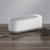 Bílý designový box nebo schránka na dřevěné podložce, vedle leží černé náramkové hodinky a brýle. Minimalistický a čistý design předmětu v neutrálních barvách.