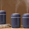 Tři tmavě šedé zvlhčovače vzduchu H2O ve stylovém designu, které současně vypouštějí jemnou páru, umístěné na dřevěném povrchu vedle knih.