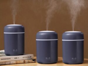 Tři tmavě šedé zvlhčovače vzduchu H2O ve stylovém designu, které současně vypouštějí jemnou páru, umístěné na dřevěném povrchu vedle knih.