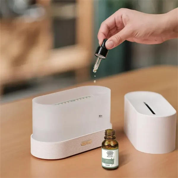 Bílý difuzér éterických olejů s lahvičkou aromaterapeutického oleje a rukou, kterákapátkem přidává několik kapek do přístroje.