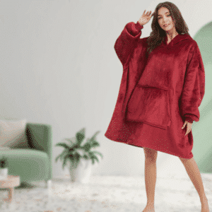 Žena v oversized červené mikině s kapucí, která vypadá pohodlně a uvolněně stojí v obývacím pokoji s rostlinou v pozadí.