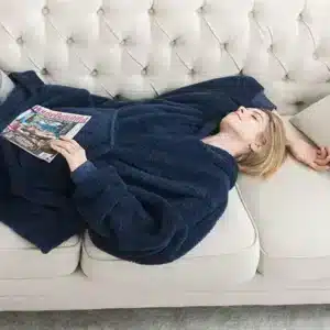Žena v modrém chlupatém oversize oblečení odpočívá na bílém čalouněném gauči a listuje časopisem, vedle ní je světlé čalounené křeslo s prošíváním.