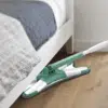 Zeleno-bílý mop s kloubovatým mopem, který čistí dřevěnou podlahu poblíž bílé postele s šedou dekou.