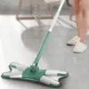 Zeleno-bílý mop s kloubovatou hlavou na úklid podlahy, v pozadí nohy osoby v domácích pantoflích.