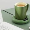 Zelený elektronický ohřívač kávy s šálkem teplého nápoje na pracovním stole vedle klávesnice, udržující konstantní teplotu nápoje.