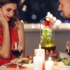 Romantická večeře při svíčkách s mladým párem, sklenicemi červeného vína a dekorativní skleněnou nádobou s osvětlenou červenou růží uprostřed stolu.