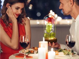 Romantická večeře při svíčkách s mladým párem, sklenicemi červeného vína a dekorativní skleněnou nádobou s osvětlenou červenou růží uprostřed stolu.