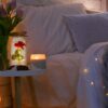 Romanticky osvětlená ložnice s kyticí fialových tulipánů, hořící svíčkou a pozlacenou růží pod skleněným poklopem na malém stolku vedle postele s jemnými světýlky.