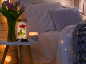 Romanticky osvětlená ložnice s kyticí fialových tulipánů, hořící svíčkou a pozlacenou růží pod skleněným poklopem na malém stolku vedle postele s jemnými světýlky.