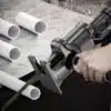 Ruční elektrická pila s pilařem řezající bílé plastové trubky na betonovém povrchu, zobrazující proces řezání potrubí.