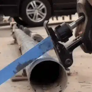 Řezačka trubek v práci, modrá rukojeť nástroje a kovová trubka na betonové podlaze s automobilovým kolem v pozadí.