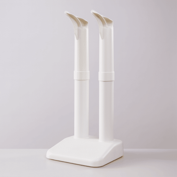 Dva bílé kosmetické štětce nebo nástroje ve vertikální poloze, umístěné na čtvercové bílé podstavě s jemným světlým pozadím.