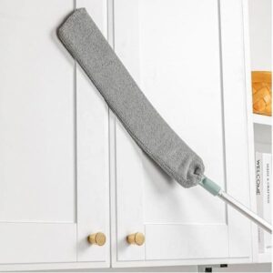 Šedý dlouhý čisticí mop s měkkým hadříkem na teleskopické tyči, zavěšený na bílé kuchyňské skříňce s mosaznou knoflíkovou klikou.