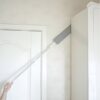 Bílá teleskopická stěrka nebo škrabka s dlouhou rukojetí dosahující na horní část bílých dveří a zárubně.