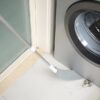 Úzká štěrbina mezi pračkou a stěnou s bílou tyčí, která brání propadnutí drobných předmětů do mezery.
