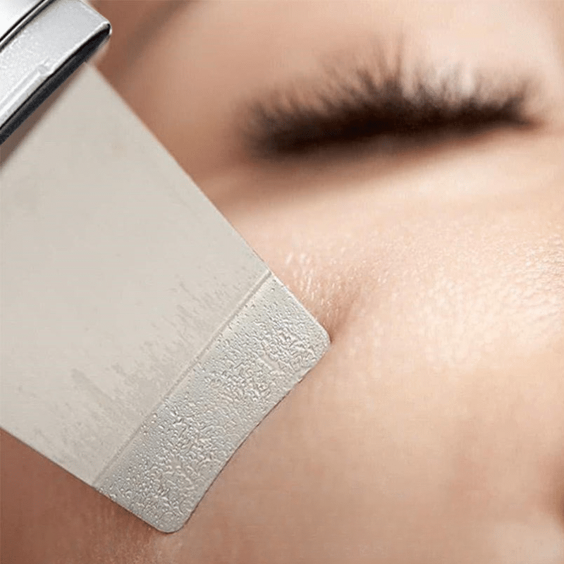 Detail mikroabrazivního nástroje těsně u lidské pokožky s rozmazaným pozadím řasy, který ukazuje postup kosmetického ošetření nebo úpravy povrchu pleti.