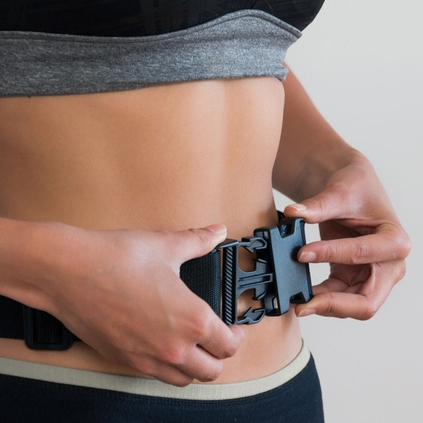 Ruce zapínající černou plastovou sponu na úzkém pásu kolem těla, který připomíná sportovní nebo fitness opasek.