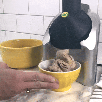 Kuchyňský přístroj na zpracování potravin, pravděpodobně robot nebo mixér, s žlutou mísou a částečně viditelnou rukou obsluhujícího.
